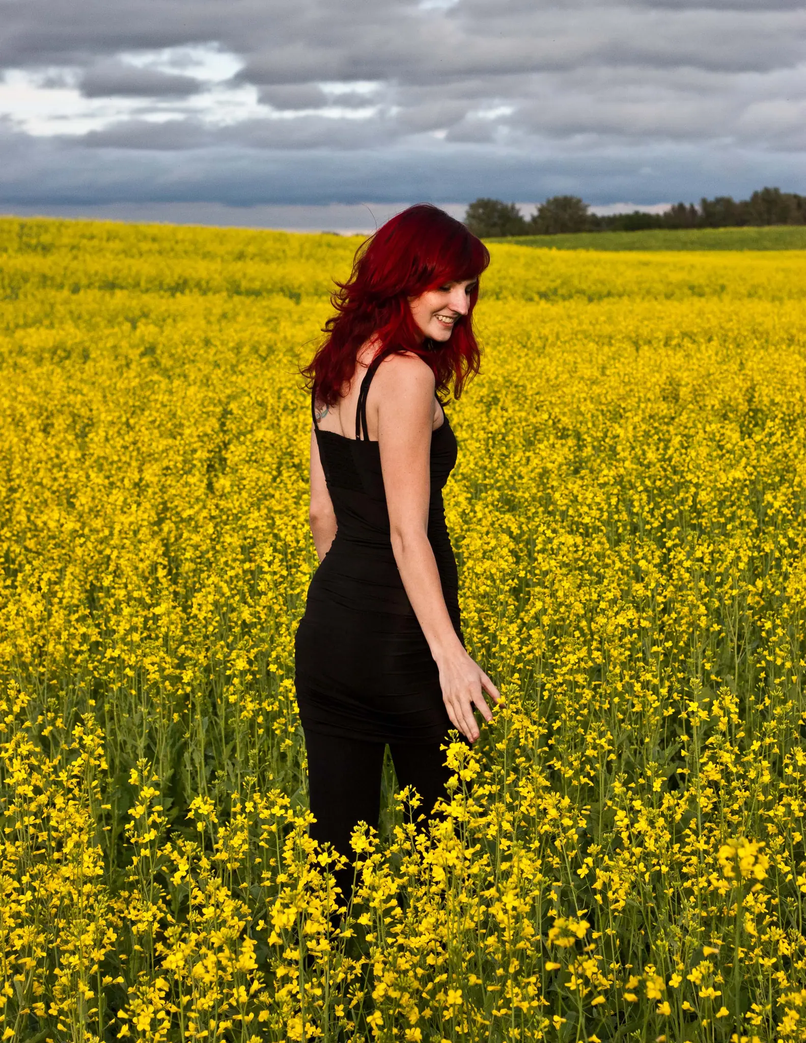 A red-haired woman standing in a canola field in full bloom under a heavy grey sky