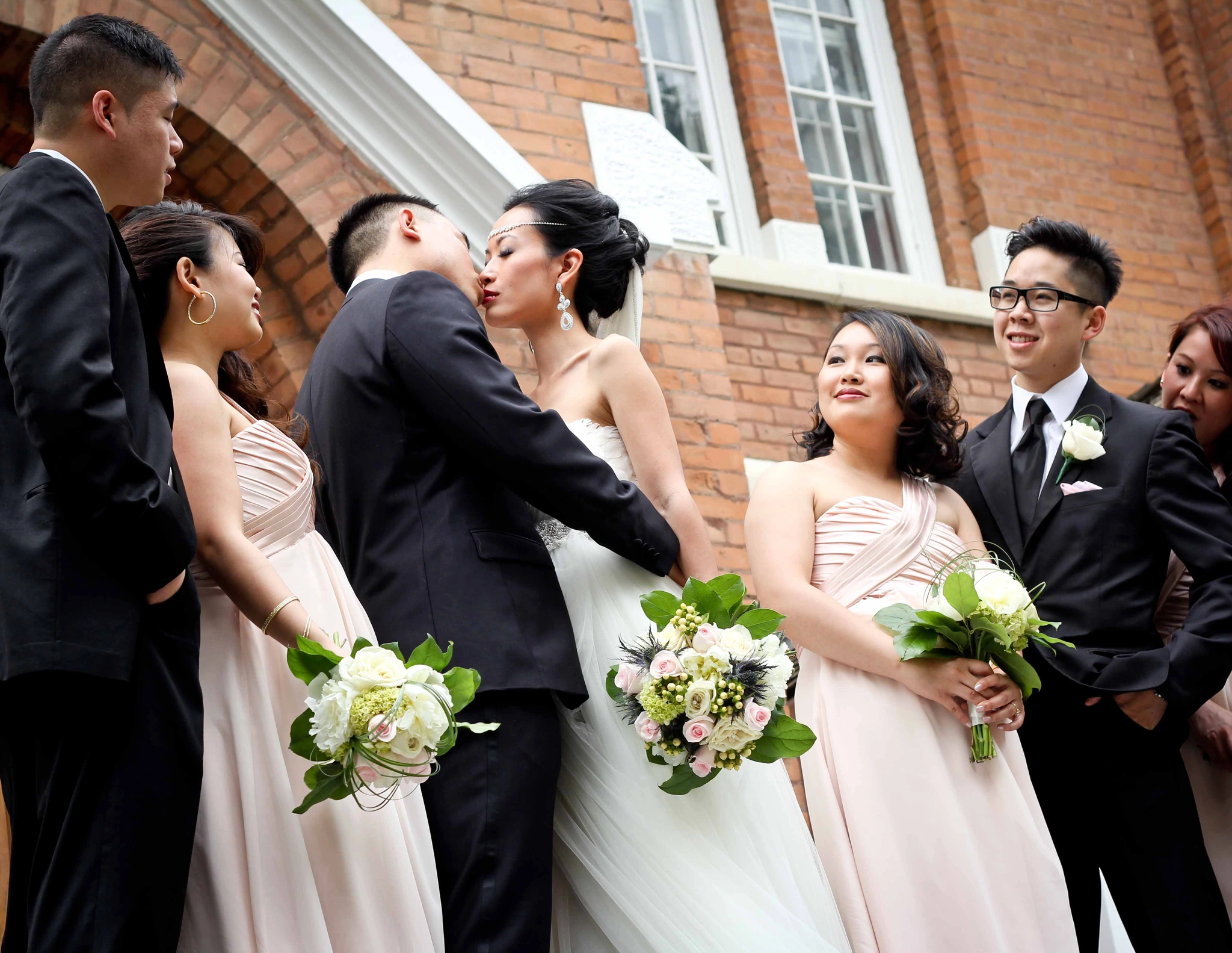 A wedding party gathered outside a brick hall, the couple kissing at its centre
